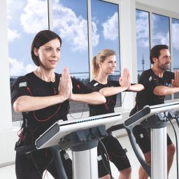 Body fit 20 minut Sp. z o.o. - Trzy osoby w strojach EMS ćwiczą w jasnym studiu z dużymi oknami. Kobieta z krótkimi włosami na pierwszym planie, za nią blondynka i mężczyzna. W tle widok na miasto.