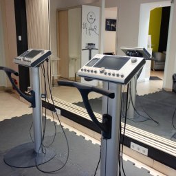 Body fit 20 minut Sp. z o.o. - Dwa urządzenia do elektrostymulacji mięśni w jasnym wnętrzu studia. Widoczne panele sterowania, uchwyty i kable. W tle lustro i elementy wyposażenia siłowni.