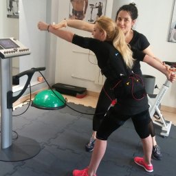 Body fit 20 minut Sp. z o.o. - Kobieta w stroju EMS ćwiczy pod okiem instruktorki w jasnym wnętrzu siłowni. Widoczny sprzęt do elektrostymulacji, rower stacjonarny i piłki do ćwiczeń.