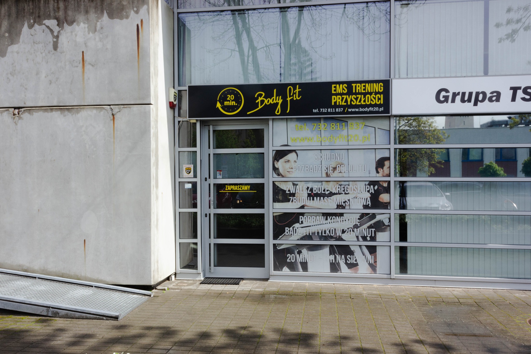 Wejście do studia Body Fit z reklamami EMS treningu: 'Schudnij, pozbądź się cellulitu, zwalcz bóle kręgosłupa'. Żółto-czarny szyld z logo i hasłem '20 minut'.