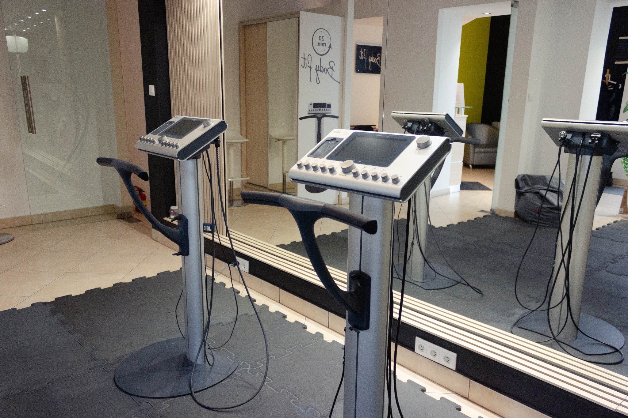 Dwa urządzenia do elektrostymulacji mięśni w jasnym wnętrzu studia. Widoczne panele sterowania, uchwyty i kable. W tle lustro i elementy wyposażenia siłowni.