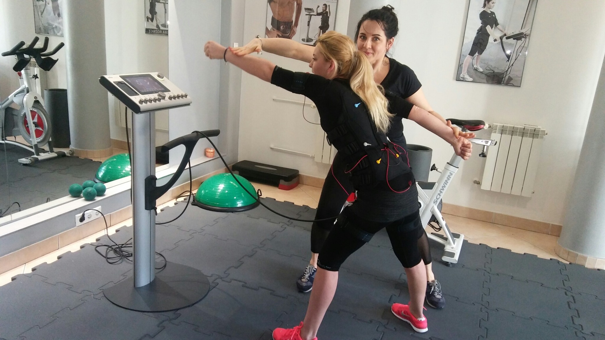 Kobieta w stroju EMS ćwiczy pod okiem instruktorki w jasnym wnętrzu siłowni. Widoczny sprzęt do elektrostymulacji, rower stacjonarny i piłki do ćwiczeń.