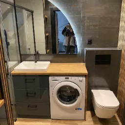 Nowoczesna łazienka z umywalką nablatową, pralką Candy, wiszącą toaletą i podświetlanym lustrem w kształcie łuku, widoczne płytki imitujące kamień i drewniane lamele.