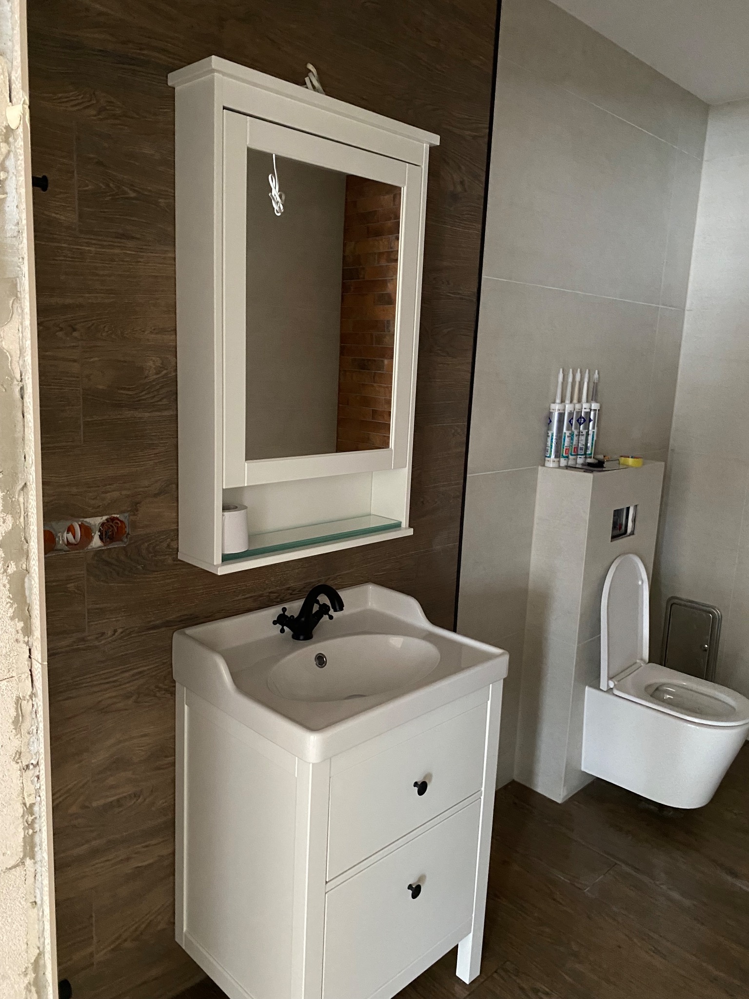 Biała szafka z umywalką i lustrem w łazience z brązowymi i szarymi płytkami. Widoczne silikony i niedokończone wykończenie ściany.