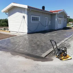 Nowo ułożona, szara kostka brukowa przed domem z szarą elewacją i czerwonym dachem, widoczna zagęszczarka wibracyjna.