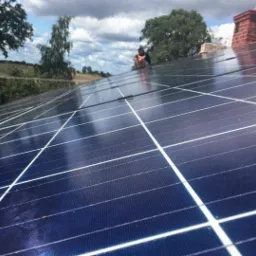 Instalacja paneli fotowoltaicznych na dachu domu z widocznym kominem z cegły i pracownikami w tle, krajobraz wiejski w oddali.