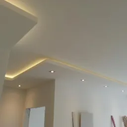 Wykończony sufit w jasnym pomieszczeniu z oświetleniem LED w listwach, widoczne punktowe światła i drabina oparta o ścianę.