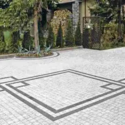 Jasnoszara kostka brukowa ułożona w geometryczny wzór kwadratu z ciemniejszymi obramowaniami, otoczona tujami i zielenią, prowadząca do domu z drewnem opałowym w tle.