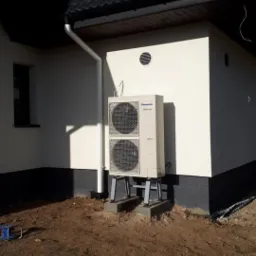 Zewnętrzna jednostka pompy ciepła Panasonic Aquarea, zamontowana na szarym stelażu na betonowych podstawach przy ścianie domu z czarnym dachem, widoczna instalacja odprowadzająca skropliny...