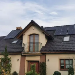Dom jednorodzinny z zainstalowanymi panelami słonecznymi na dachu pokrytym ciemną dachówką, widoczne okna dachowe i balkon.
