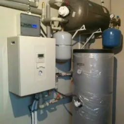 Kompleksowa instalacja grzewcza w kotłowni: zbiornik akumulacyjny owinięty folią, bojler, ekspansomat i rozbudowany system rur.