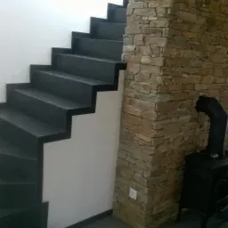 Ciemne schody z kamiennymi stopniami i wykończeniem ściany z kamienia naturalnego w odcieniach beżu i szarości, obok stoi czarny piec.