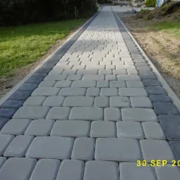 Ścieżka z jasnoszarej kostki brukowej z ciemnoszarym obramowaniem, widok perspektywiczny w kierunku trawnika i hałdy ziemi.