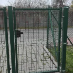 Zielona, metalowa furtka z siatki z czarnym zamkiem, zamontowana na betonowej podmurówce, widok na szarą kostkę brukową i nagie drzewa w tle.