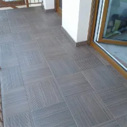 Wyłożony płytkami taras w odcieniach szarości i brązu, z wzorem szachownicy utworzonym przez ułożenie płytek z różnym wzorem słojów drewna, widok z boku balkonu z drewnianą balustradą i oknem.