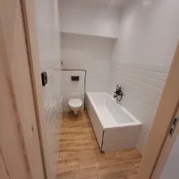 Nowoczesna łazienka z białymi płytkami typu metro, drewnopodobną podłogą, białą wanną z czarną armaturą i podwieszaną toaletą z zabudowanym systemem spłukiwania.