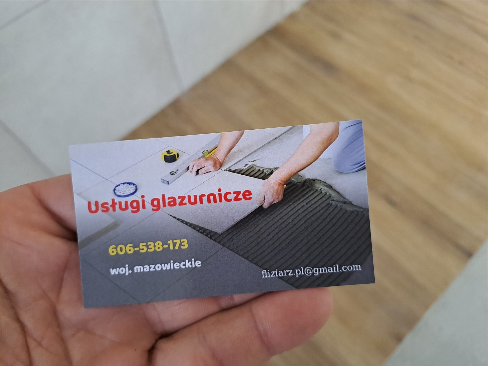 W dłoni wizytówka z grafiką przedstawiającą układanie płytek, numerem telefonu i adresem email.