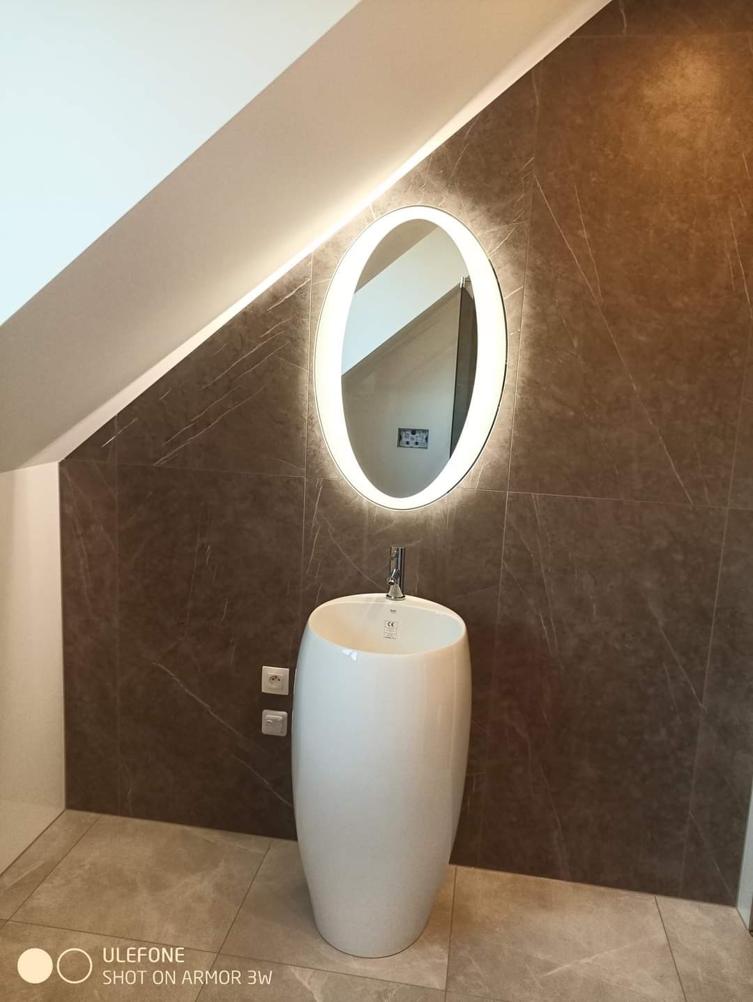 Nowoczesna łazienka na poddaszu z umywalką wolnostojącą i podświetlanym lustrem owalnym, brązowe płytki imitujące marmur na ścianie, szare płytki podłogowe.