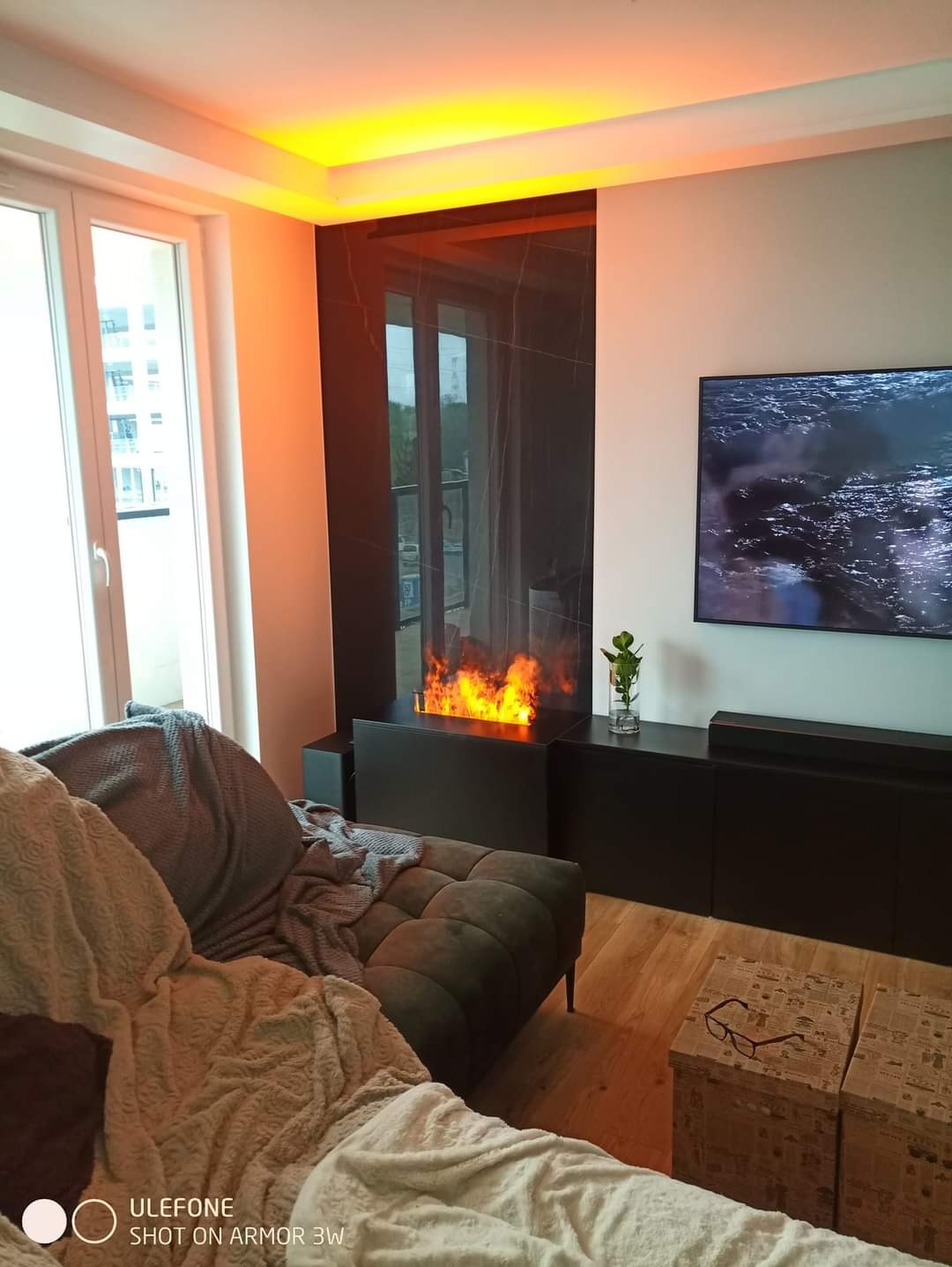 Salon z biokominkiem w zabudowie z czarnego marmuru, podświetlony sufit, szara sofa z narzutą i telewizor z widokiem na morze.