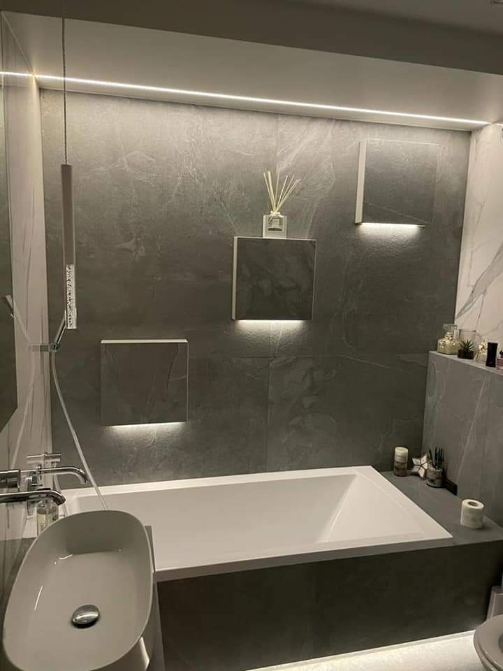 Nowoczesna łazienka z szarymi płytkami imitującymi kamień, podświetlanymi kwadratowymi panelami ściennymi, białą wanną i umywalką nablatową, minimalistyczna aranżacja z akcentami dekoracyjnymi.