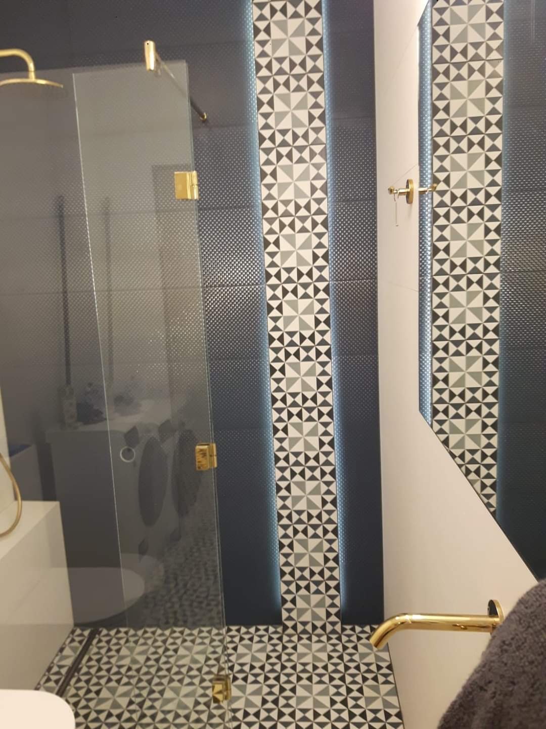 Nowoczesna łazienka z kabiną prysznicową ze złotymi okuciami, ściany wyłożone ciemnymi płytkami z delikatnym wzorem oraz pasami dekoracyjnymi z geometrycznym motywem trójkątów w odcieniach szarości...