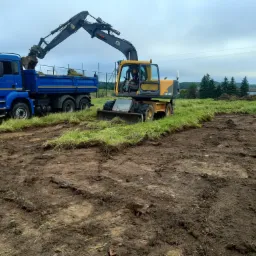 Żółty koparko-ładowarka z operatorem nabiera ziemię na niebieską ciężarówkę MAN na terenie budowy, widoczne ślady opon na ziemi i trawa w tle.