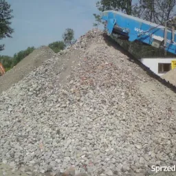 Góra gruzu i kruszywa z niebieskim podajnikiem taśmowym w tle, widoczny fragment budynku i drzewa.