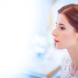 M Pro Studio. Fotograf - Portret z profilu panny młodej z rudymi włosami upiętymi w kok, subtelny makijaż, perłowe kolczyki i naszyjnik, delikatna koronka sukni ślubnej i fragment welonu.