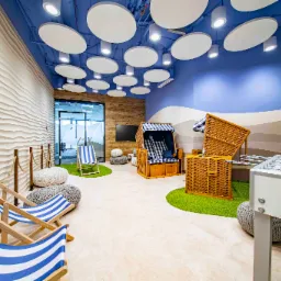 Kreatywna aranżacja biura z motywem plażowym: leżaki w niebiesko-białe paski, wiklinowe kosze plażowe, stół do piłkarzyków, sztuczna trawa i falista ściana w tle, dopełnione okrągłymi lampami...