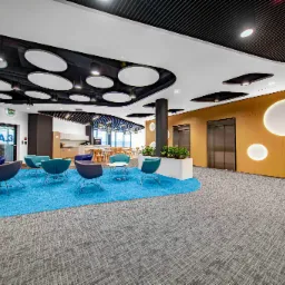 Nowoczesna przestrzeń biurowa z niebieskim dywanem, krzesłami w różnych odcieniach, oświetleniem sufitowym w kształcie dysków i drewnianą ścianą z okrągłymi podświetleniami.