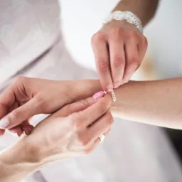 Zapinanie srebrnej bransoletki na nadgarstku panny młodej z delikatnym, koronkowym mankietem, widoczne dłonie z różowym manicure i pierścionek zaręczynowy.
