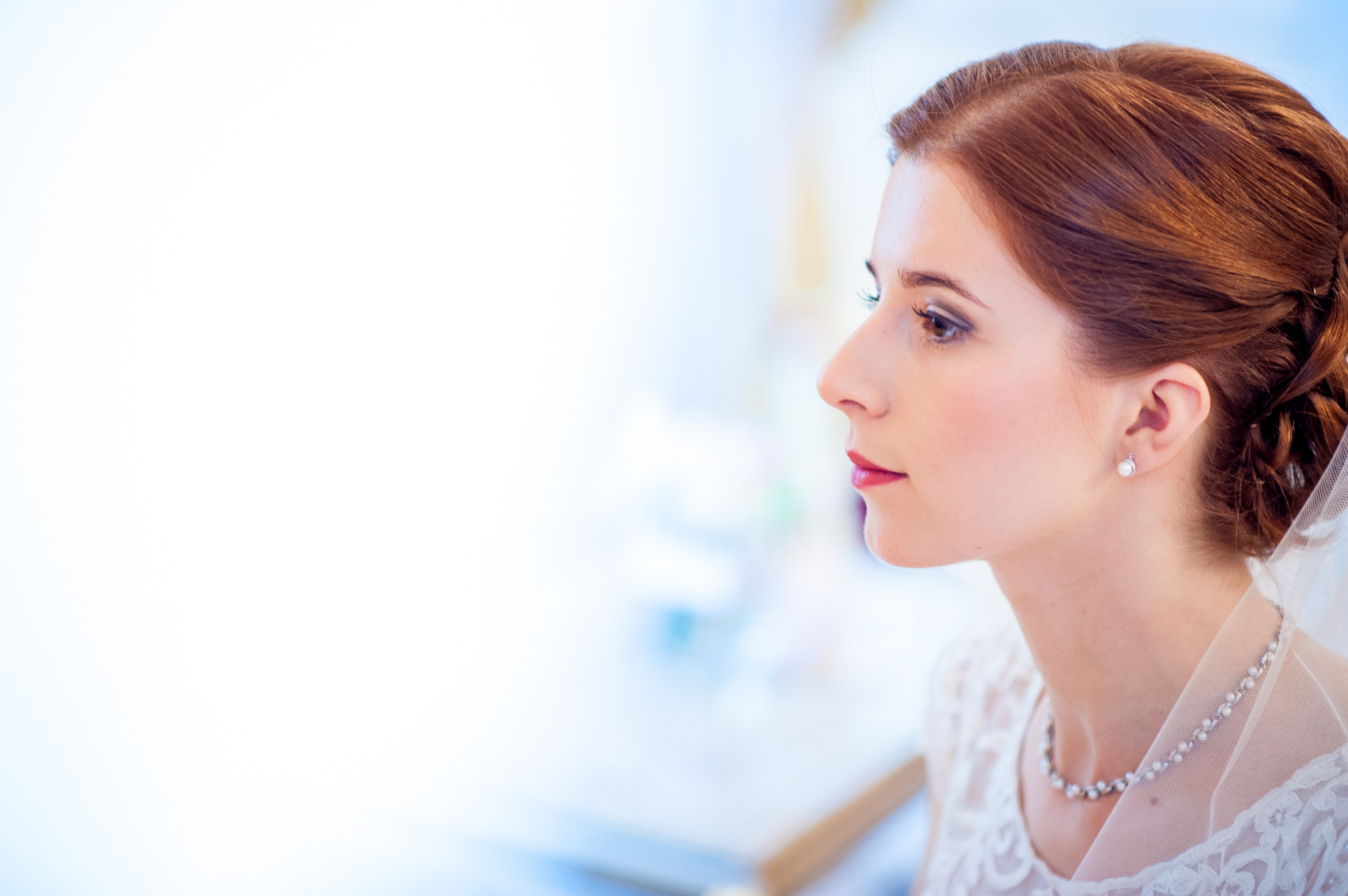 Portret z profilu panny młodej z rudymi włosami upiętymi w kok, subtelny makijaż, perłowe kolczyki i naszyjnik, delikatna koronka sukni ślubnej i fragment welonu.