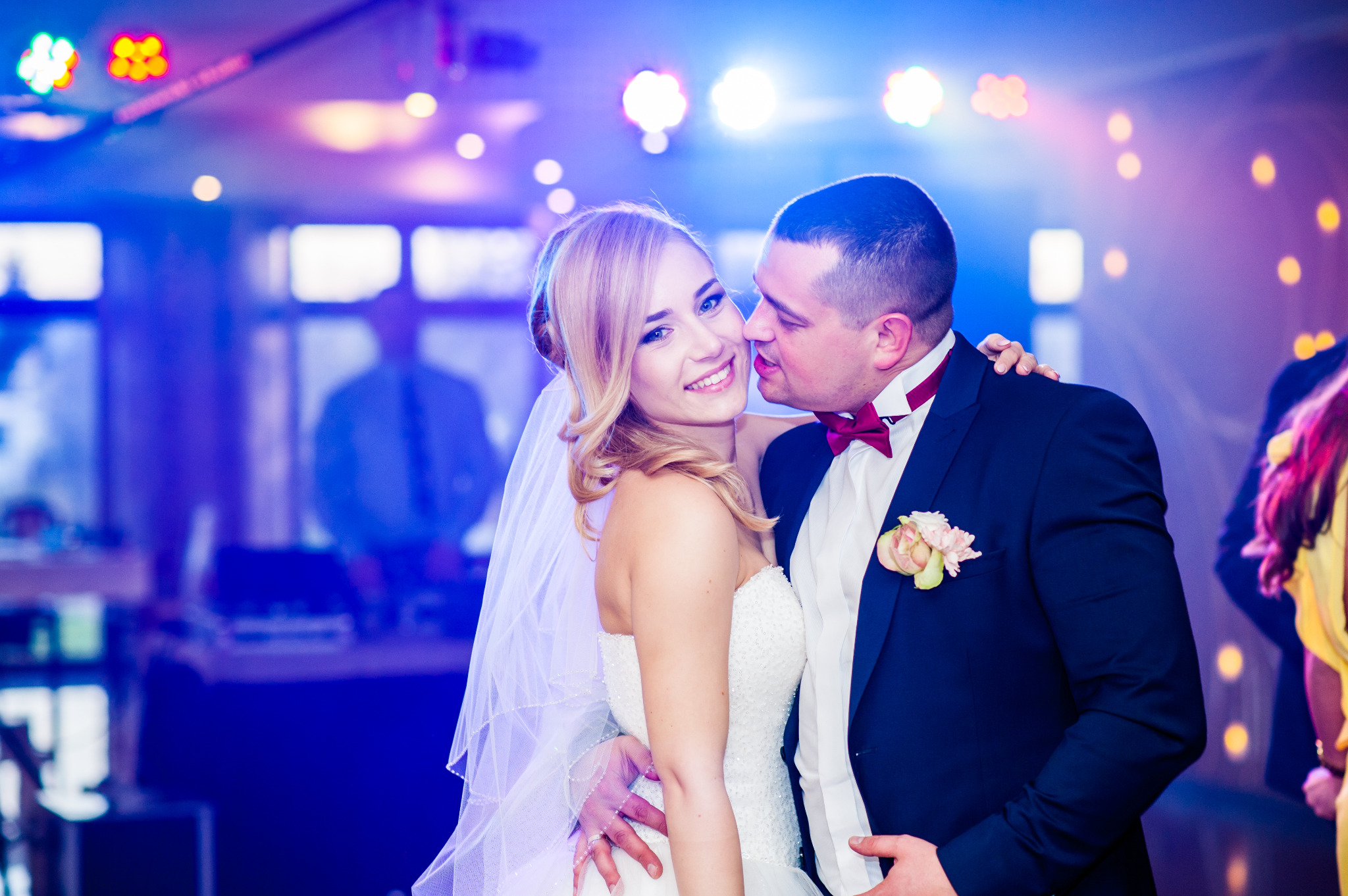 Uśmiechnięta panna młoda w białej sukni i welonie, obejmowana przez pana młodego w granatowym garniturze z bordową muchą, na tle sceny z oświetleniem dyskotekowym.