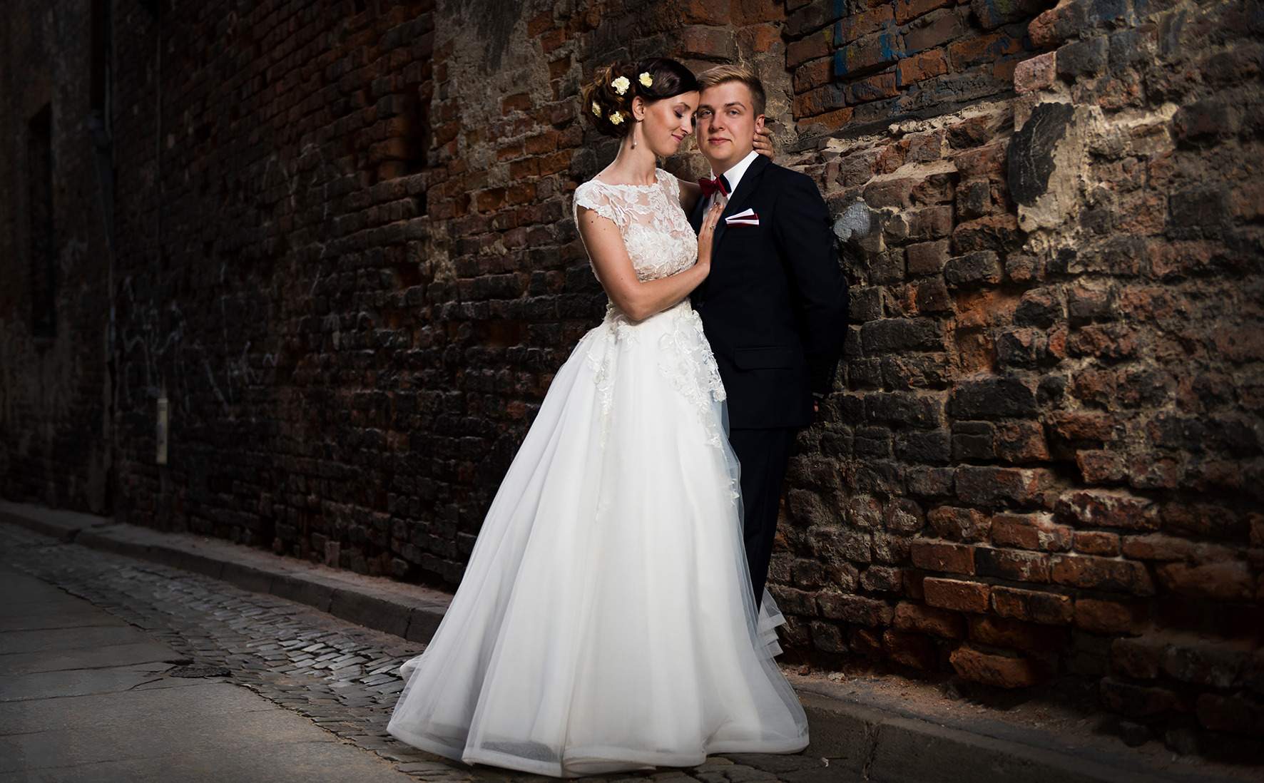 Para młoda w eleganckich strojach pozuje na tle starej ceglanej ściany na brukowanej uliczce, panna młoda ma wpięte kwiaty we włosy, a pan młody czerwoną muchę i poszetkę.