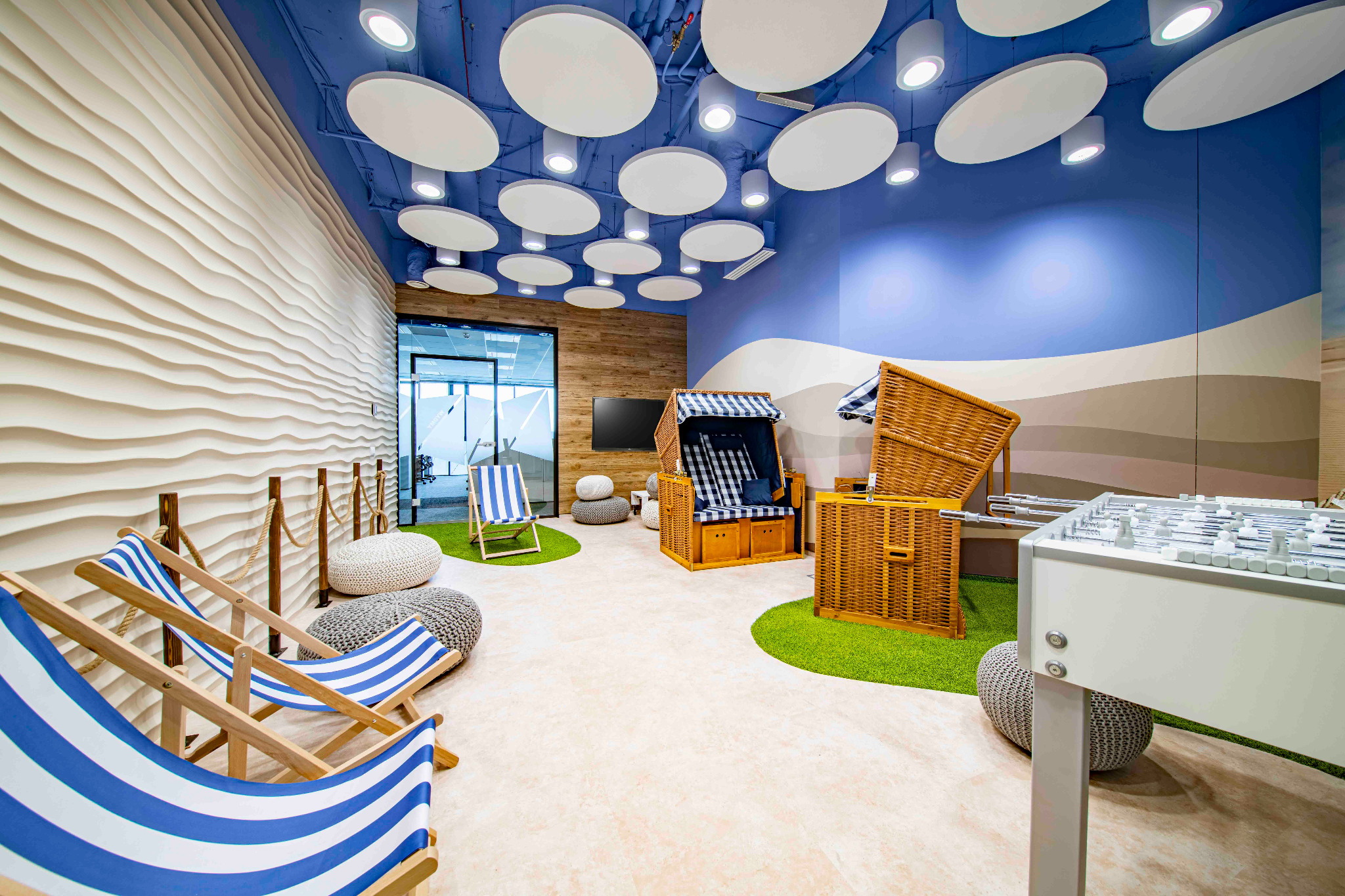 Kreatywna aranżacja biura z motywem plażowym: leżaki w niebiesko-białe paski, wiklinowe kosze plażowe, stół do piłkarzyków, sztuczna trawa i falista ściana w tle, dopełnione okrągłymi lampami...