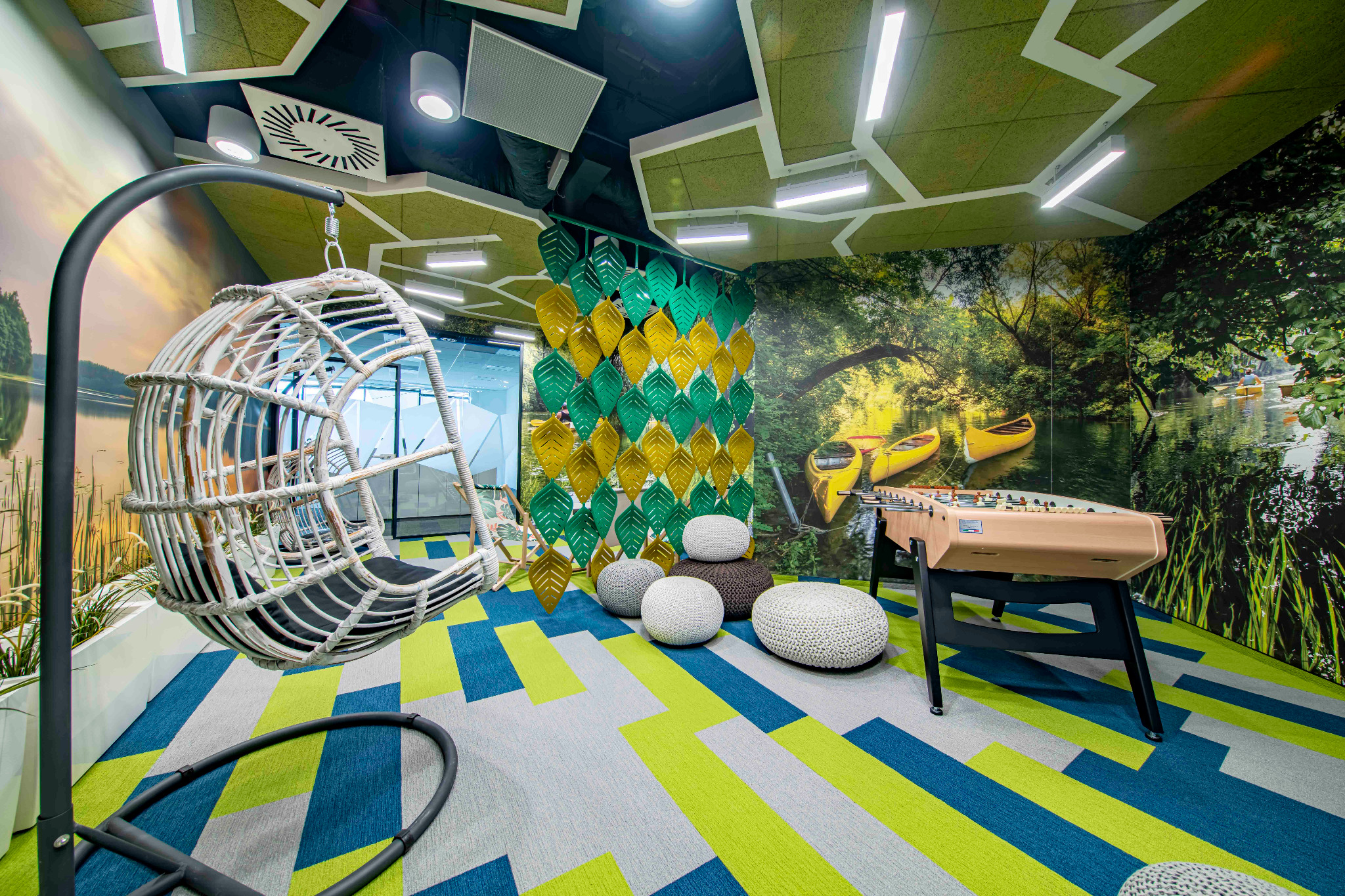 Nowoczesna przestrzeń biurowa z huśtawką kokon, stołem do piłkarzyków, pufami i fototapetą z motywem kajaków na rzece, charakteryzująca się geometrycznym wzorem na podłodze i dekoracyjnym panelem...