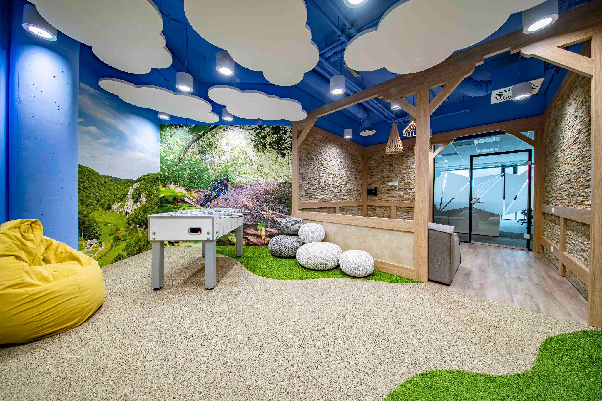 Kreatywna aranżacja pokoju relaksu z motywem natury: fototapeta leśna, sufit w chmurki, piłkarzyki, pufy i drewniana konstrukcja.