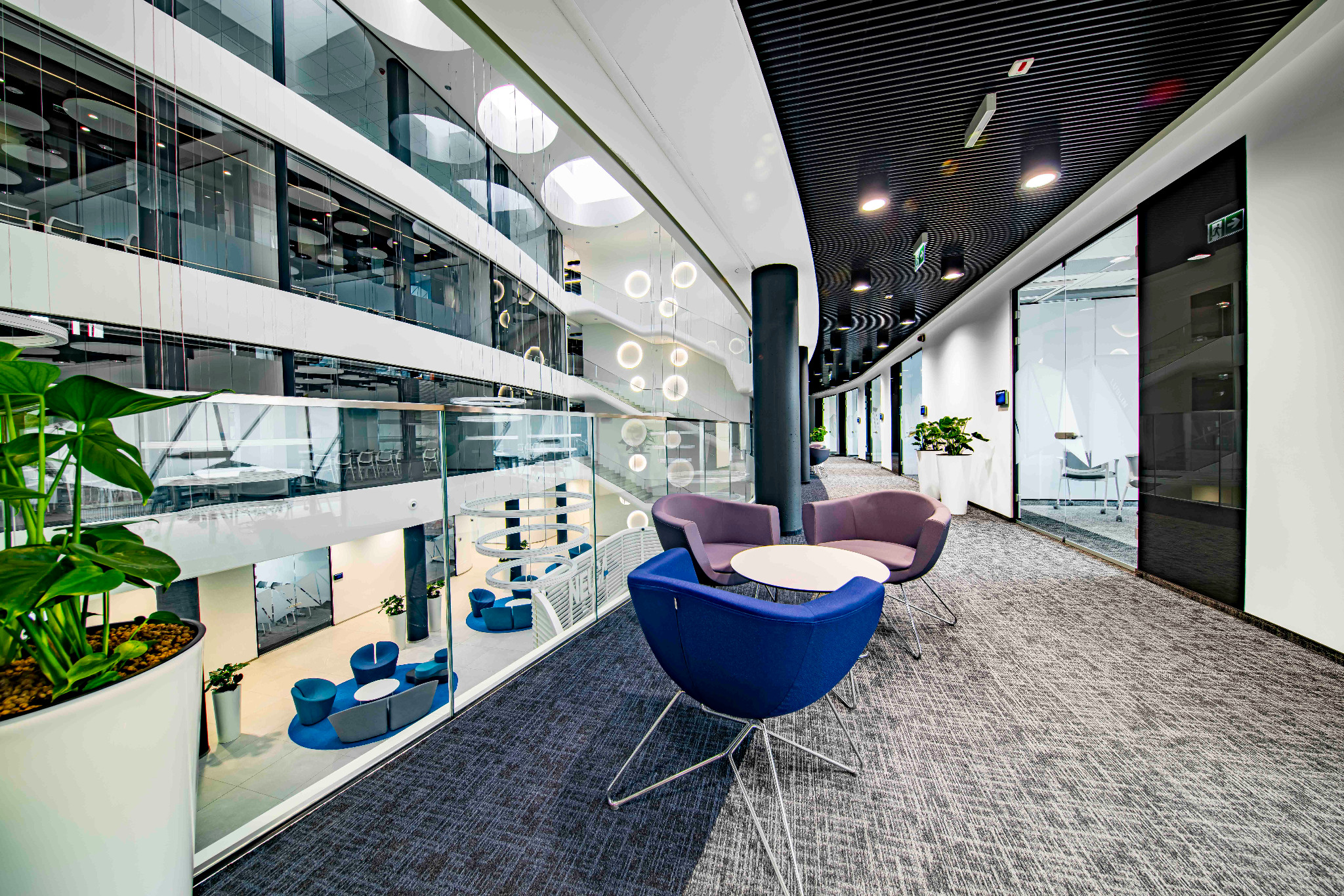 Wnętrze biura z nowoczesnym designem, widok z antresoli na strefę relaksu z kolorowymi fotelami i stolikiem, na tle przeszklonej ściany widoczny hol z okrągłymi lampami.