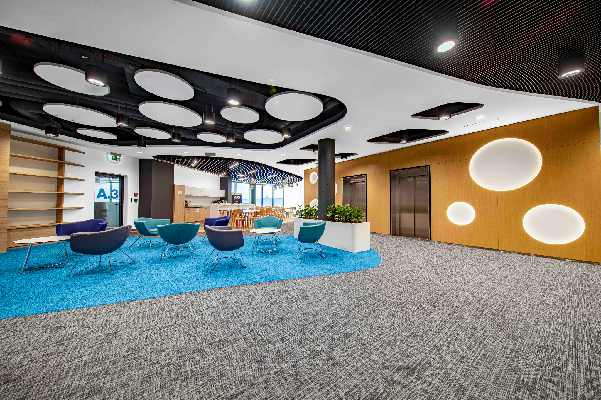 Nowoczesna przestrzeń biurowa z niebieskim dywanem, krzesłami w różnych odcieniach, oświetleniem sufitowym w kształcie dysków i drewnianą ścianą z okrągłymi podświetleniami.