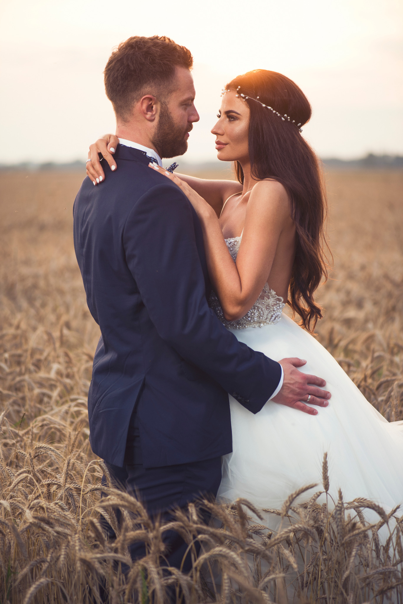 Para młoda obejmująca się na polu zboża w promieniach zachodzącego słońca; panna młoda w sukni bez ramiączek z ozdobnym paskiem i wiankiem, pan młody w granatowym garniturze z muszką.