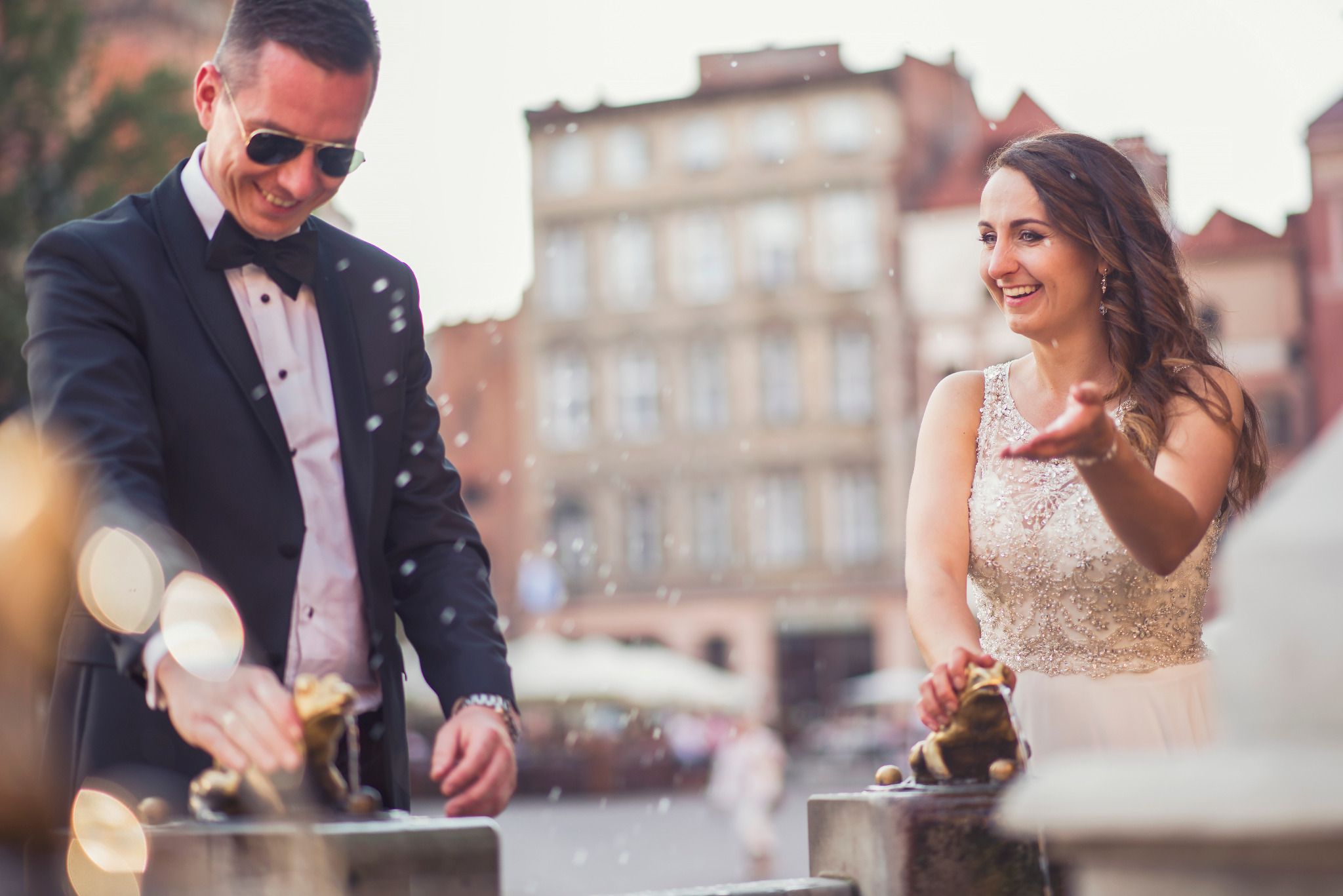 Uśmiechnięta para młoda w eleganckich strojach, dotyka fontanny z figurkami żab w historycznym centrum miasta, w tle zabytkowe kamienice i delikatny efekt bokeh.
