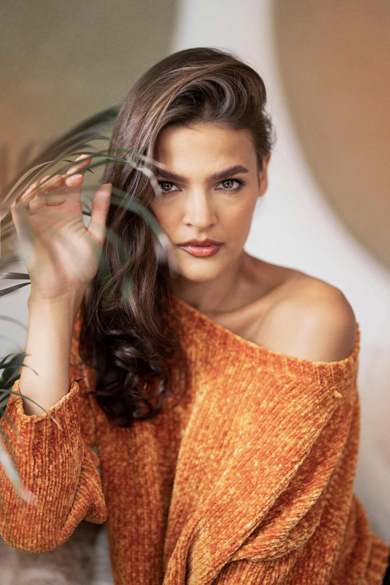 Portret kobiety w pomarańczowym swetrze z odkrytym ramieniem, pozującej z zielonym liściem na pierwszym planie, wzrok skierowany w obiektyw.