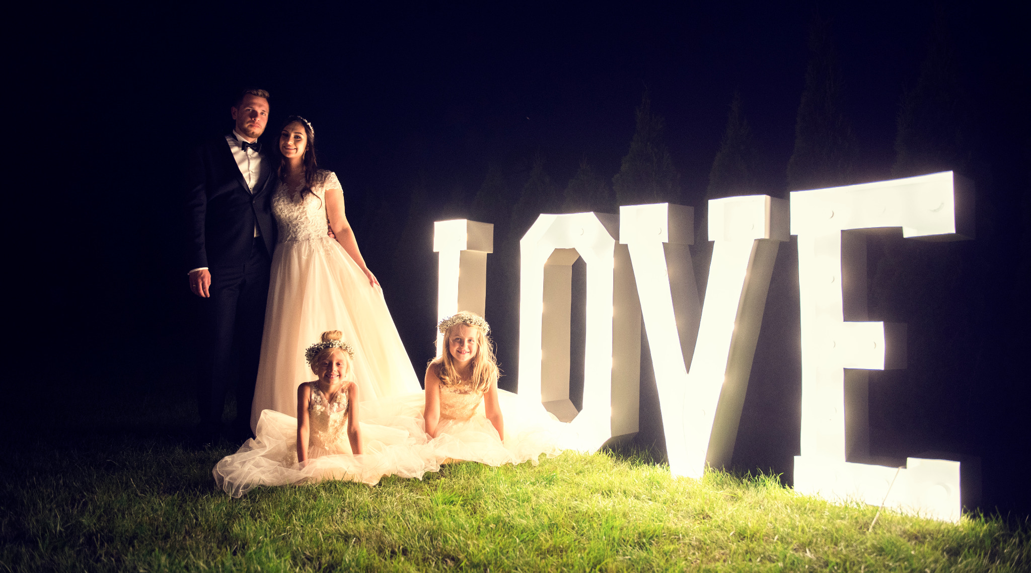 Para młoda z dwiema dziewczynkami w sukienkach, pozuje na trawie w Papowo Toruńskie, obok dużego, podświetlanego napisu LOVE, na tle ciemnego nieba.