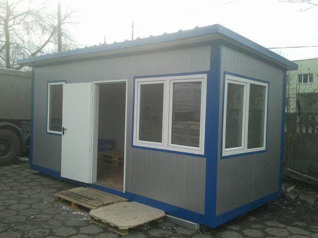 Gotowa stróżówka z płyty warstwowej w kolorze szarym z niebieskimi obramowaniami, z otwartymi drzwiami i widocznymi paletami w środku, ustawiona na zewnątrz na kostce brukowej, obok zaparkowany...