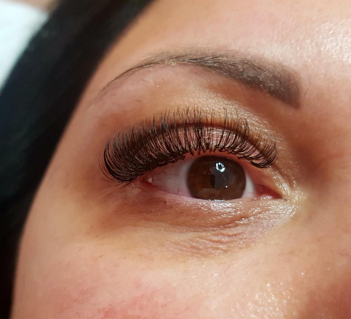 Zbliżenie na oko z brązową tęczówką i gęstymi, profesjonalnie przedłużonymi rzęsami w stylu 'kocie oko', widoczny delikatny makijaż brwi.