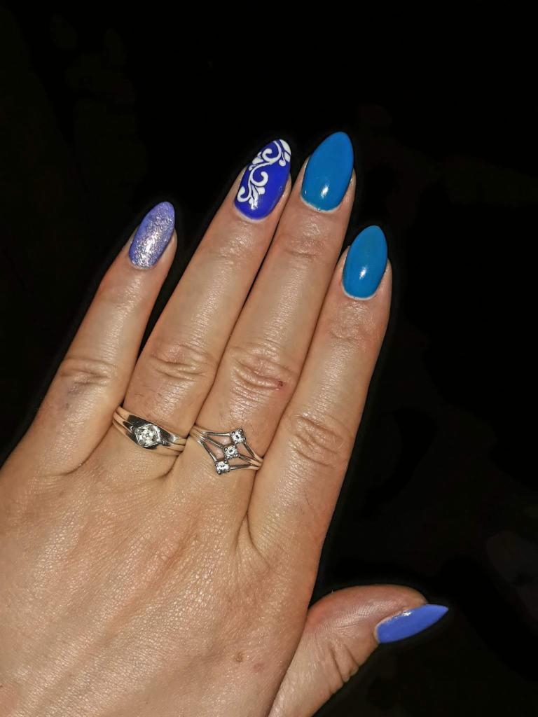 Dłoń z manicure hybrydowym: jeden paznokieć brokatowy, dwa turkusowe, jeden z białym ornamentem na kobaltowym tle, dwa srebrne pierścionki.