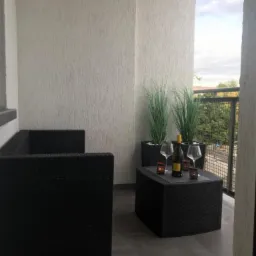 Balkon z czarnymi meblami rattanowymi, butelką wina i kieliszkami na stoliku, wykończony tynkiem strukturalnym.