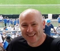 Uśmiechnięty mężczyzna w czarnej koszulce na tle trybun stadionu, z widoczną zieloną murawą w oddali.