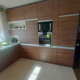 Nowoczesna zabudowa kuchenna z drewnopodobnymi frontami, srebrnymi listwami, wbudowanym piekarnikiem i mikrofalówką, oraz otwartymi półkami z dekoracjami.