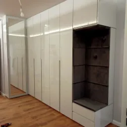 Biała szafa wnękowa z połyskiem, zintegrowana z tapicerowanym siedziskiem i oświetleniem LED, widoczna częściowo w lustrze, z wkrętarką na drewnianej podłodze.